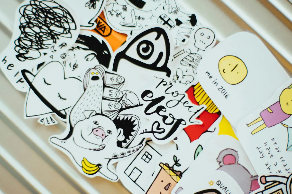A pile of hand-drawn doodle stickers, most die-cut to their own outline with a thin white border and a few cut as rounded rectangles, spread across a ridged metal surface