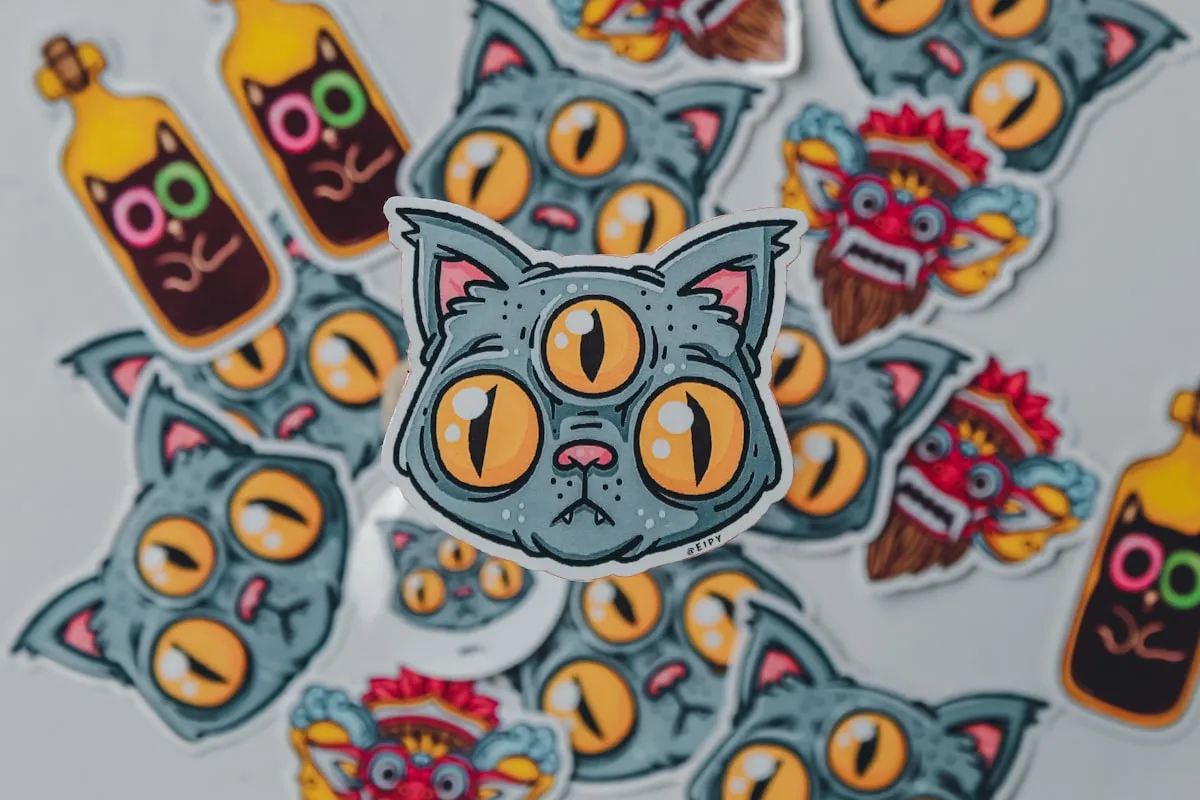 A scattered flat-lay of colorful die-cut vinyl stickers — a three-eyed cat, tribal masks, and potion bottles — spread across a light surface