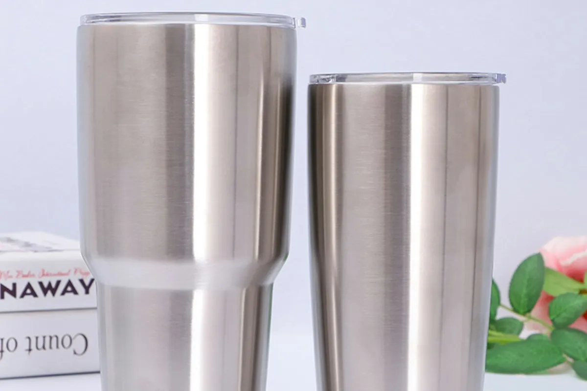 Two blank stainless steel insulated tumblers with clear lids standing side by side on a light surface, the kind of polymer-coated blanks used for sublimation printing