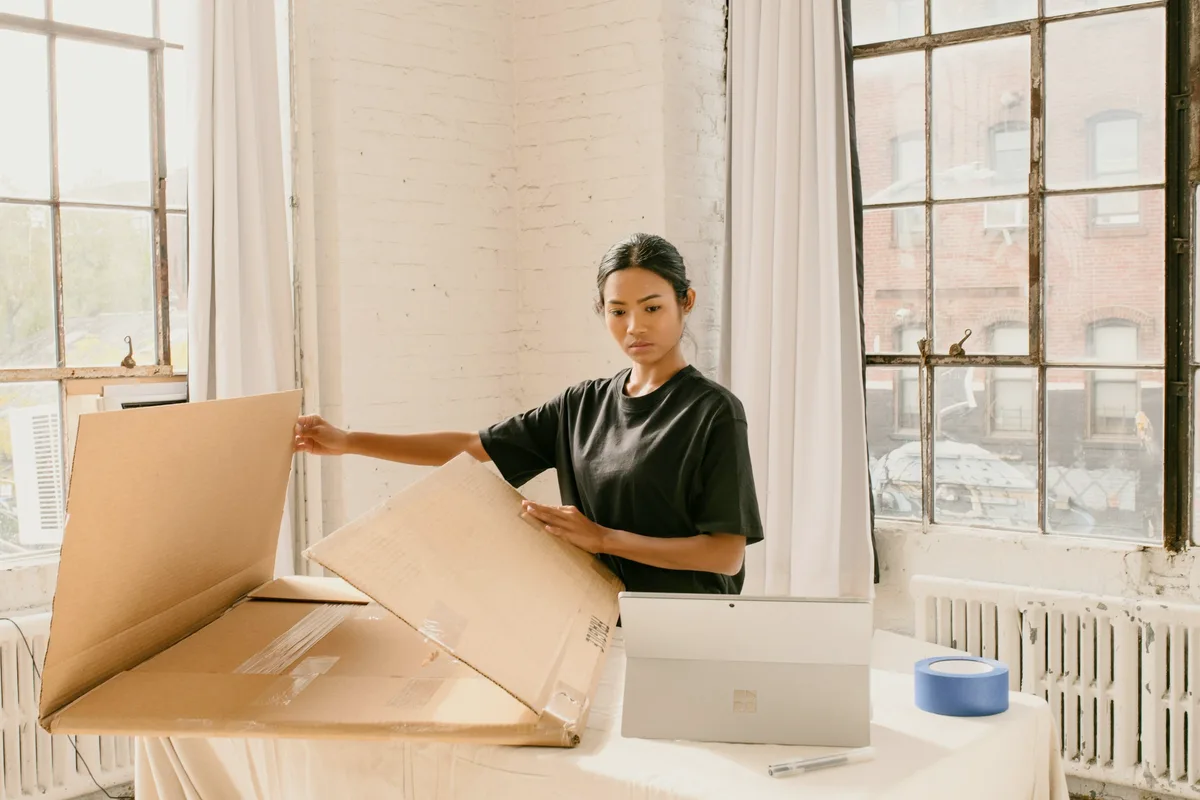 A woman in a black t-shirt folding open a large flat cardboard shipping box on a cloth-covered table beside an open laptop and a roll of blue tape, in a bright loft room with tall windows