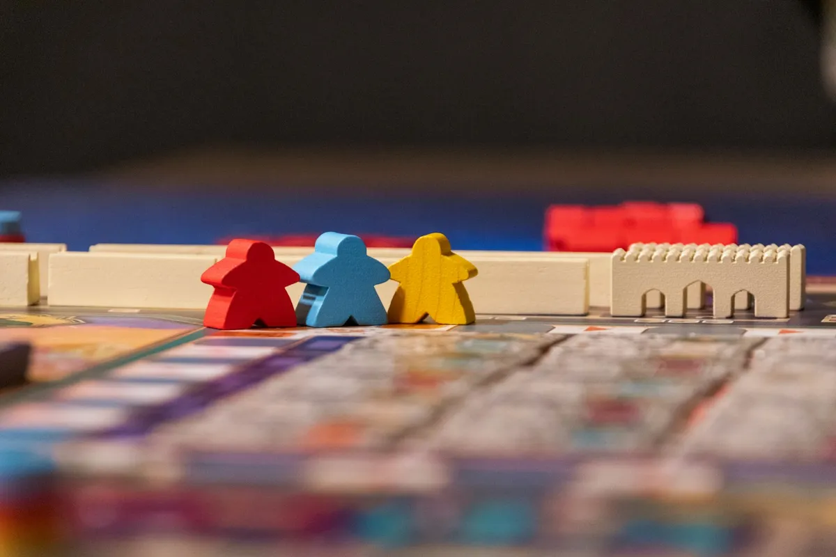 Three wooden meeples — red, blue, and yellow — standing on a colorful indie tabletop game board next to a small wooden castle piece