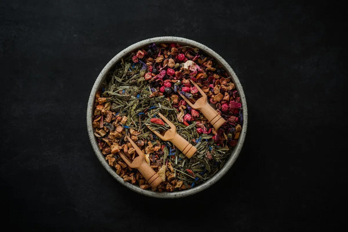 Overhead view of a stone bowl of loose herbal tea blend with green leaves, pink petals, red berries and blue cornflowers, and three wooden tea scoops resting in it