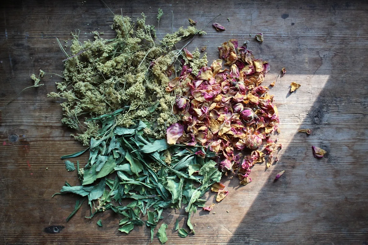 Piles of dried botanicals — green herb leaves, pink and gold rose petals, and seed heads — sorted on a rustic dark wood table for hand-blending tea