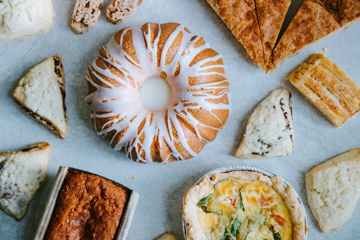 An overhead flat-lay of home-baked goods on a pale gray surface — a glazed lemon bundt cake at center, a vegetable quiche in a foil tin, a loaf of banana bread, biscotti, sliced cinnamon scones, and fruit-filled pastry triangles — spanning the non-TCS and TCS food categories now authorized under the Tennessee Food Freedom Act