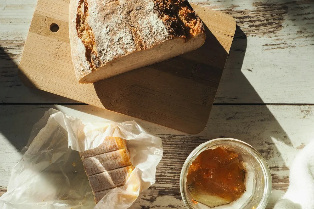 A rustic sourdough loaf rests on a wooden cutting board next to a glass jar of orange marmalade and a bundle of crackers wrapped in parchment, on weathered whitewashed farmhouse boards in afternoon sunlight