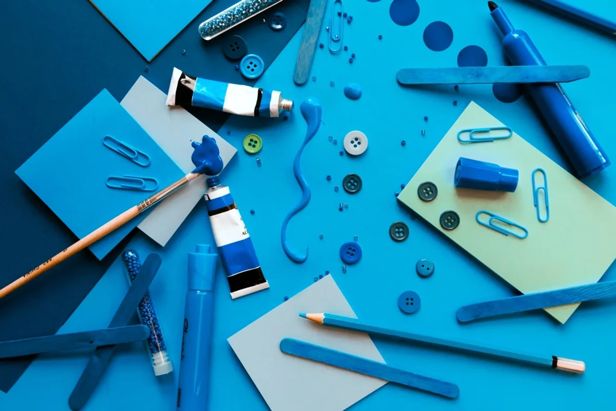An overhead flat-lay of arts and crafts supplies — pens, paint, buttons, paper clips, and pencils — arranged on a blue background, evoking a maker's materials rather than a retail shelf