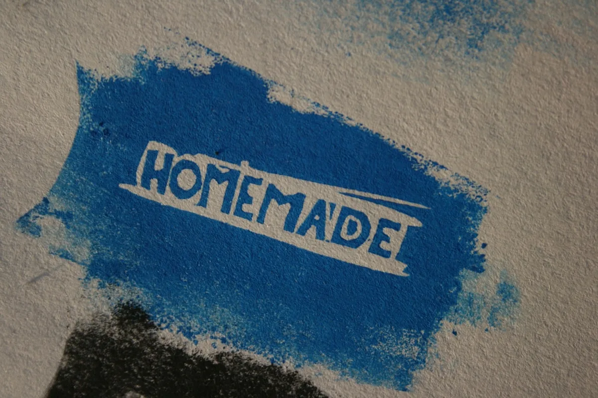 Close-up of the word HOMEMADE hand-stamped in blue ink across a rough patch of paint on a pale textured wall, with a smudged black mark below it