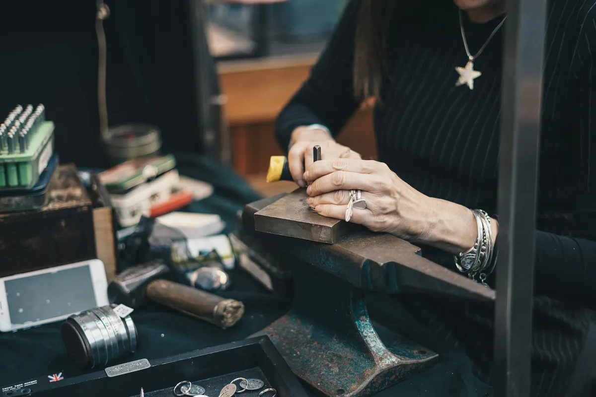 A jeweler in a dark sweater working at a cluttered metalsmithing bench, hands holding a stamp tool over a steel block and anvil with scattered findings, files, and small parts in the foreground