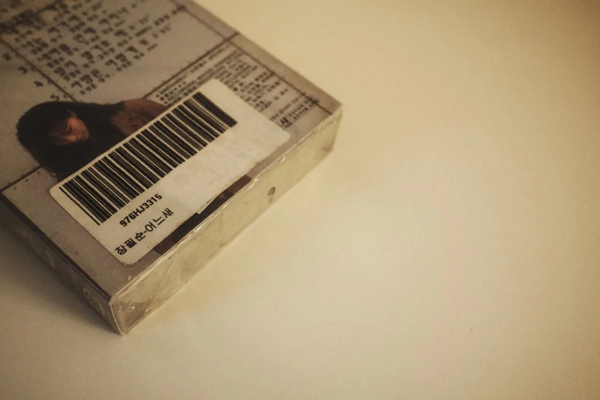 A worn white barcode sticker on the corner of a clear plastic product case, its printed insert showing small photo artwork and text, photographed at an angle on a cream surface with open space to the right