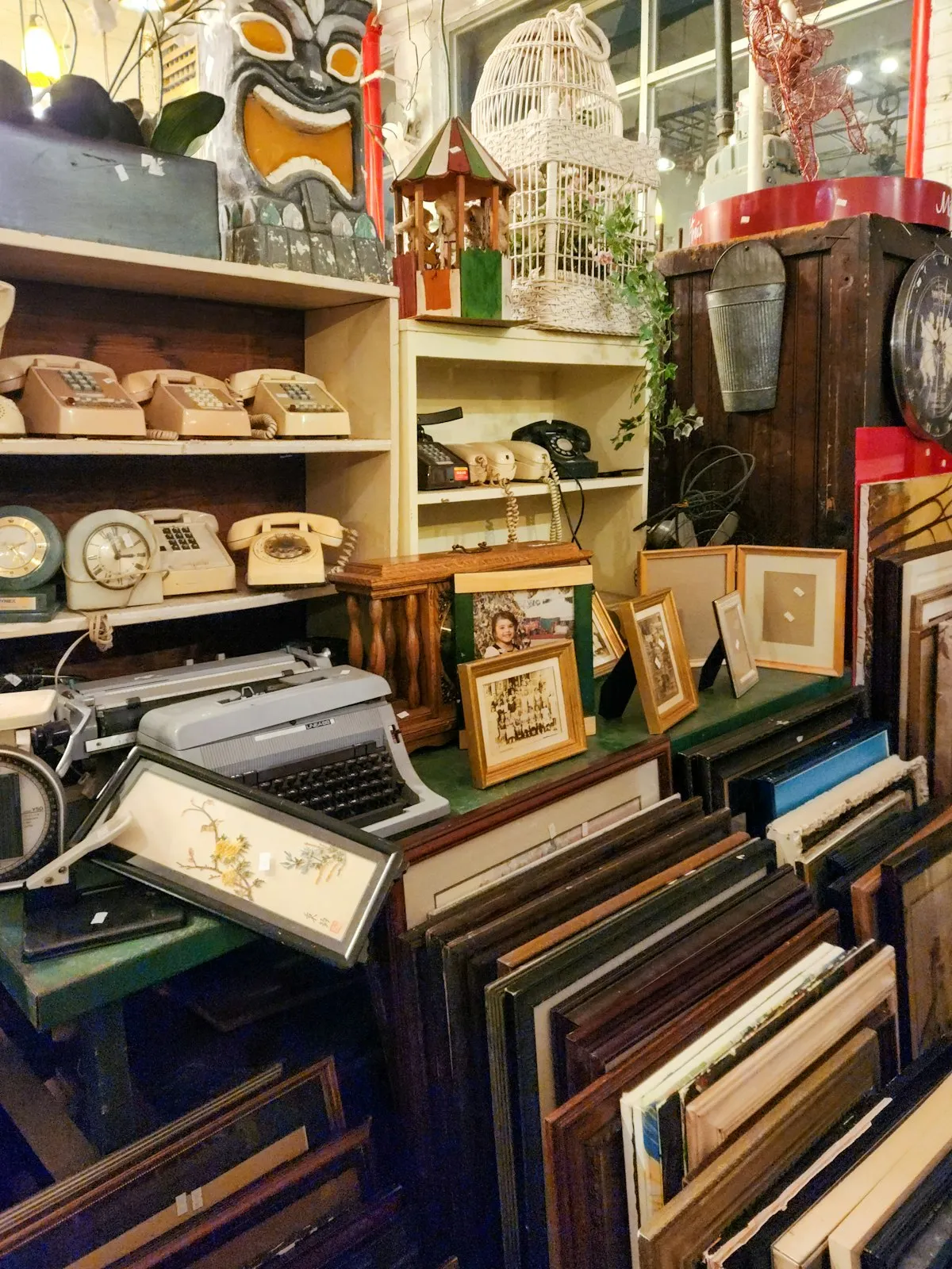 Cluttered antique shop interior with vintage telephones, a typewriter, framed artwork, a birdcage, and assorted curiosities on wooden shelves