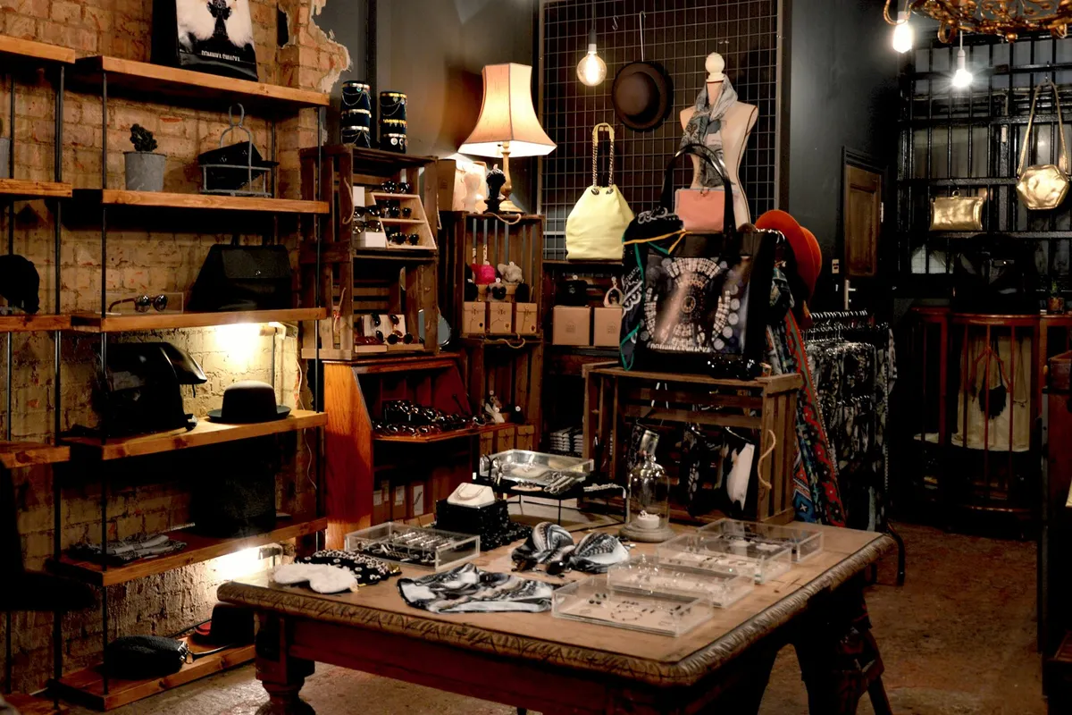 Interior of a warmly lit vintage boutique with rustic wooden crates holding sunglasses, hats, jewelry, and small accessories on open shelves, hanging handbags on a mannequin, and a low jeweler's display table covered in necklaces and trinkets in the foreground