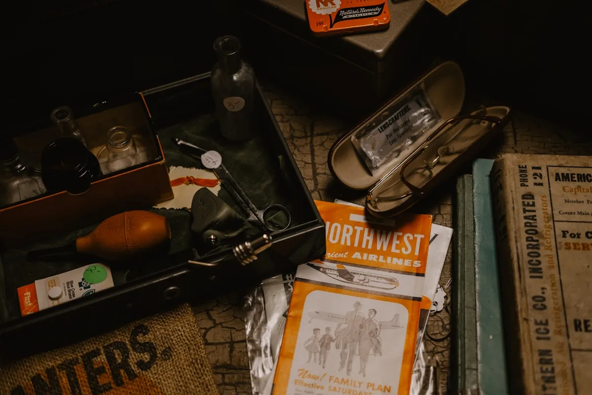 A moody flat lay of estate-sale finds — an open vintage toolbox with glass bottles, antique eyeglasses in a case, a 1950s airline travel brochure, an old hardcover ledger, and a tin of mint cachous arranged on a textured tabletop