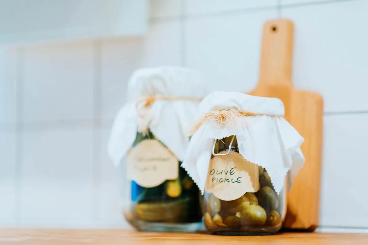 Two glass jars of home-pickled vegetables sealed with cloth covers tied in twine, each with a handwritten paper label reading "Olive Pickle" and "Cucumber Pickle", sitting on a wooden countertop against a sunlit white-tile kitchen backsplash