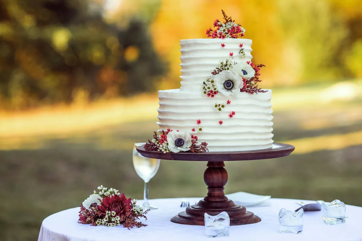 A two-tier white textured buttercream wedding cake topped with fresh flowers on a wooden cake stand at an outdoor venue table, with warm autumn foliage blurred behind it