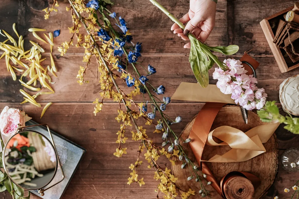 Overhead flat-lay of a florist's wooden worktable mid-arrangement — a hand holding a stem against branches of yellow forsythia, blue delphinium, pale pink stock and roses, with copper ribbon, kraft paper, a small round mirror and a wood disc scattered across the surface
