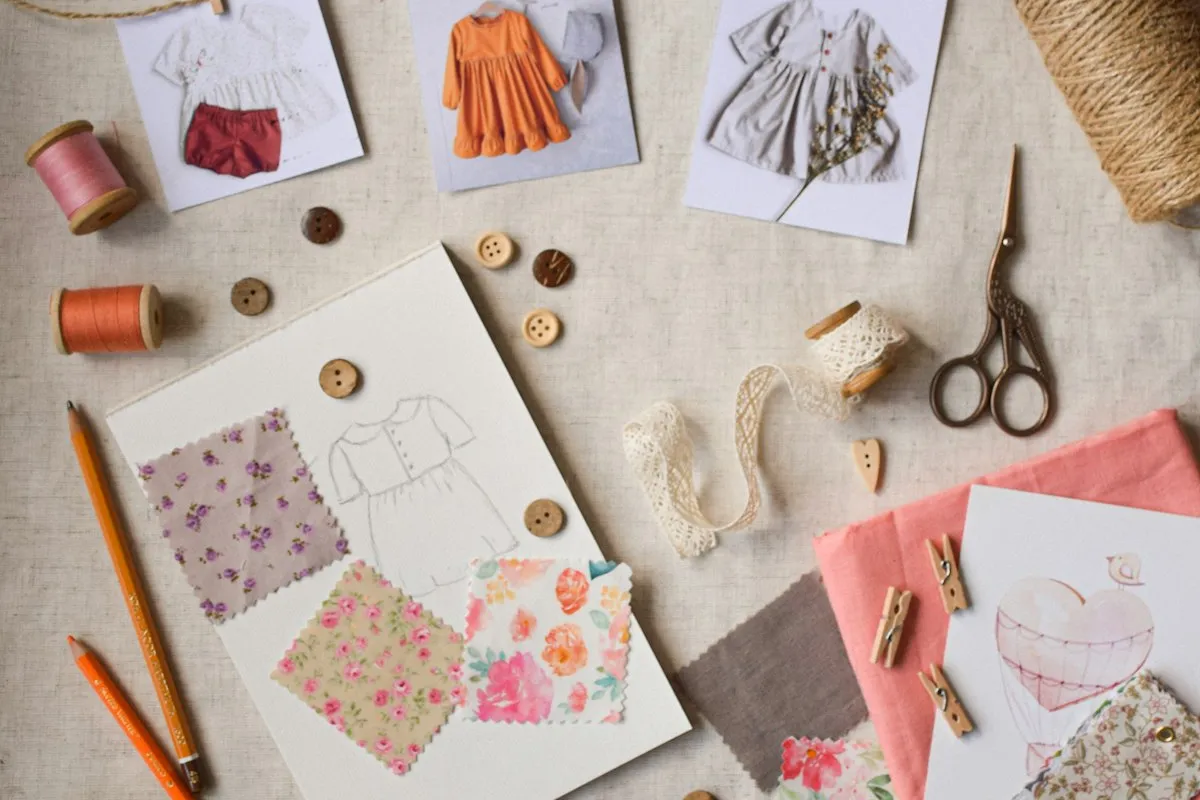 An overhead flat lay of a garment sewing project — a dress sketch surrounded by fabric swatches, wooden buttons, thread spools, lace trim, embroidery scissors, and pencils on a linen surface
