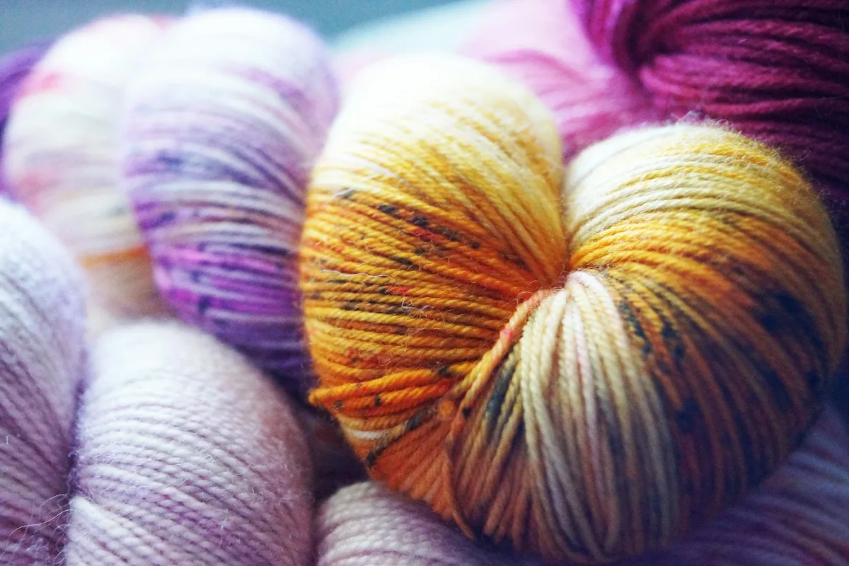 Close-up of twisted skeins of hand-dyed yarn: a golden-orange speckled skein at the center, with a speckled lilac skein and pale pink skeins to its left and below and a deep magenta skein behind it at the upper right, each with uneven, hand-applied color