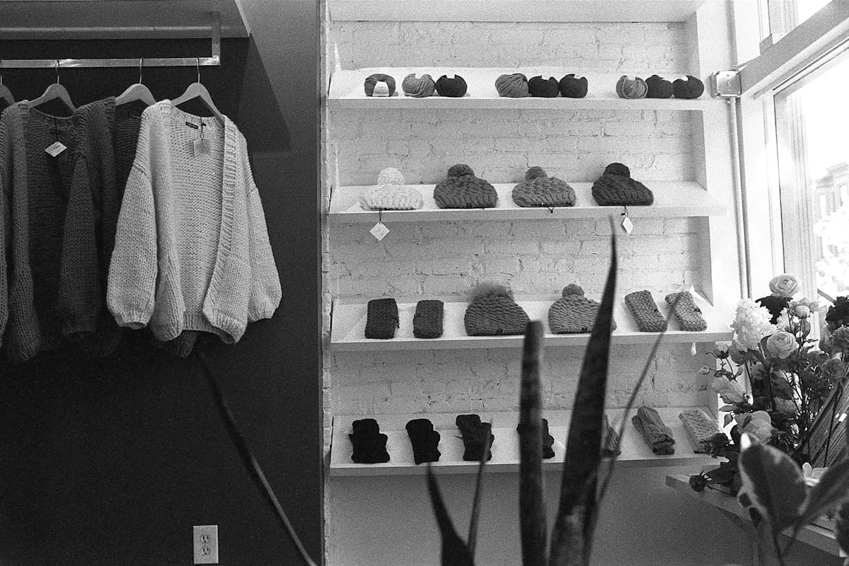 A black-and-white photo of a small boutique interior — handmade chunky-knit hats and balls of yarn arranged across four white-brick wall shelves, with hand-knit sweaters hanging on a rack to the left and a window with flowers and a snake plant to the right