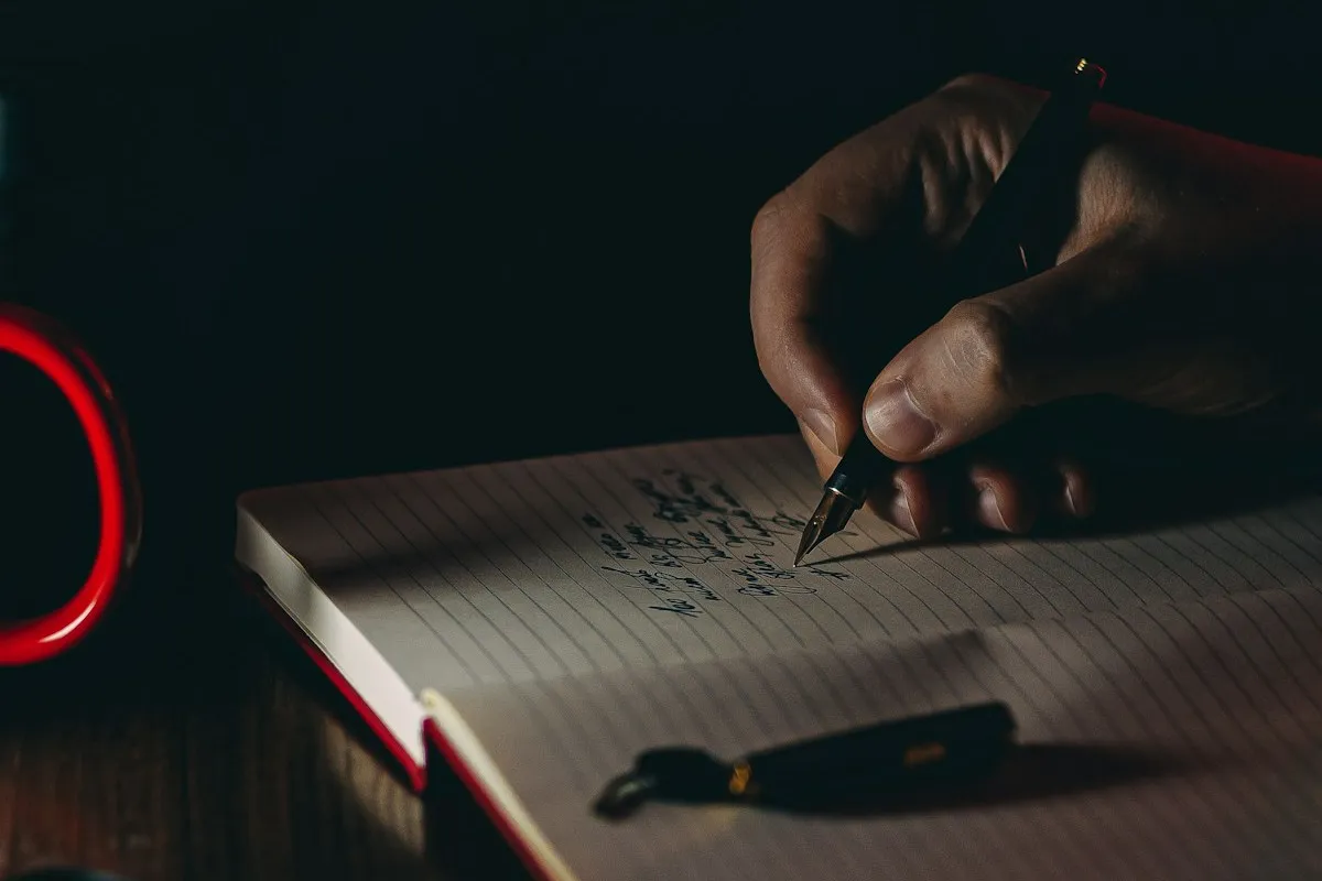A hand writing in a lined notebook by lamplight at a dark desk, a fountain pen cap resting on the open page