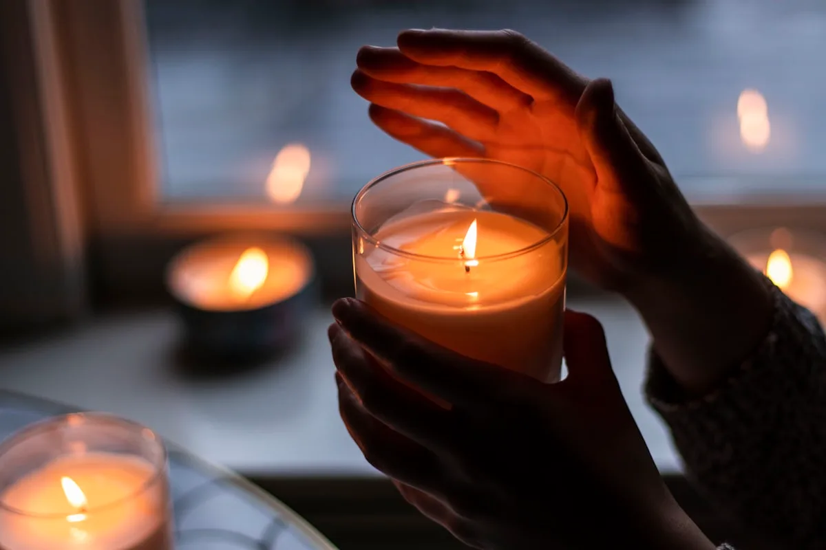 Hands cupped around a lit candle in a clear glass tumbler on a windowsill at dusk, with more candles burning nearby and cold blue light in the window behind — the drafty, cold-glass conditions a wick with no margin will fail in