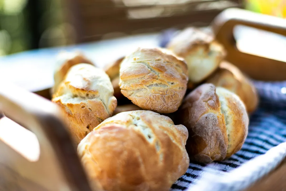 A wooden tray of golden, crusty home-baked dinner rolls resting on a blue-and-white striped tea towel, photographed in soft natural light with a soft-focus garden background