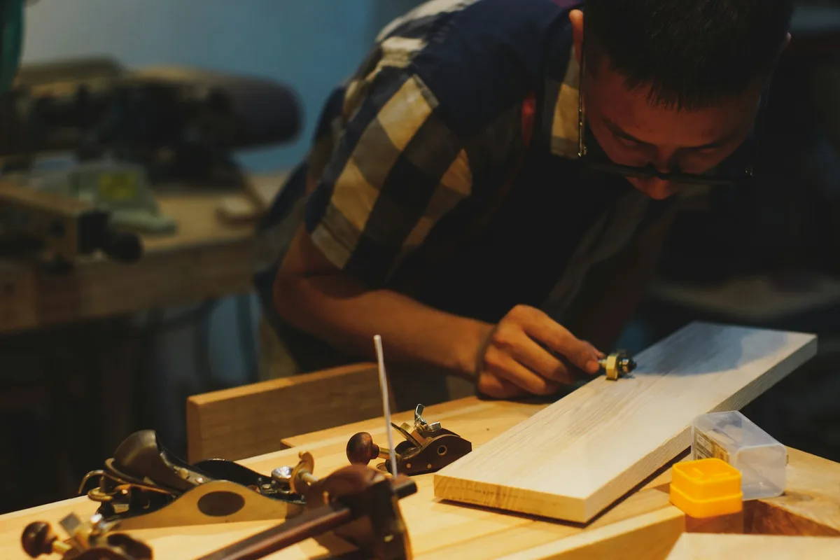 A woodworker leaning close over a planed board, marking it with a gauge, with two hand planes and a marking tool resting on the bench in the foreground of a warmly lit shop