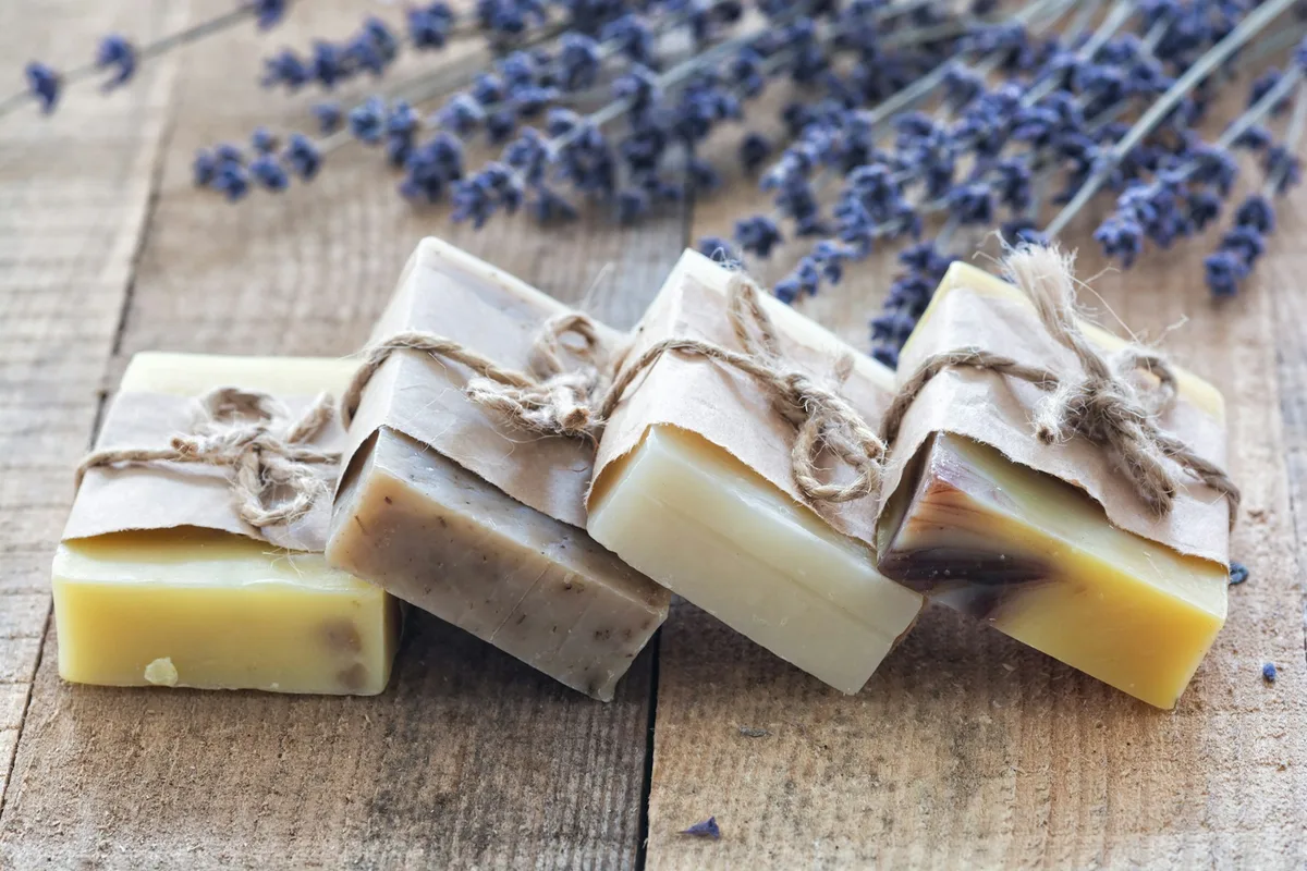 Four bars of handmade soap, each wrapped in a paper band tied with twine, lined up on a weathered wooden board in front of dried lavender