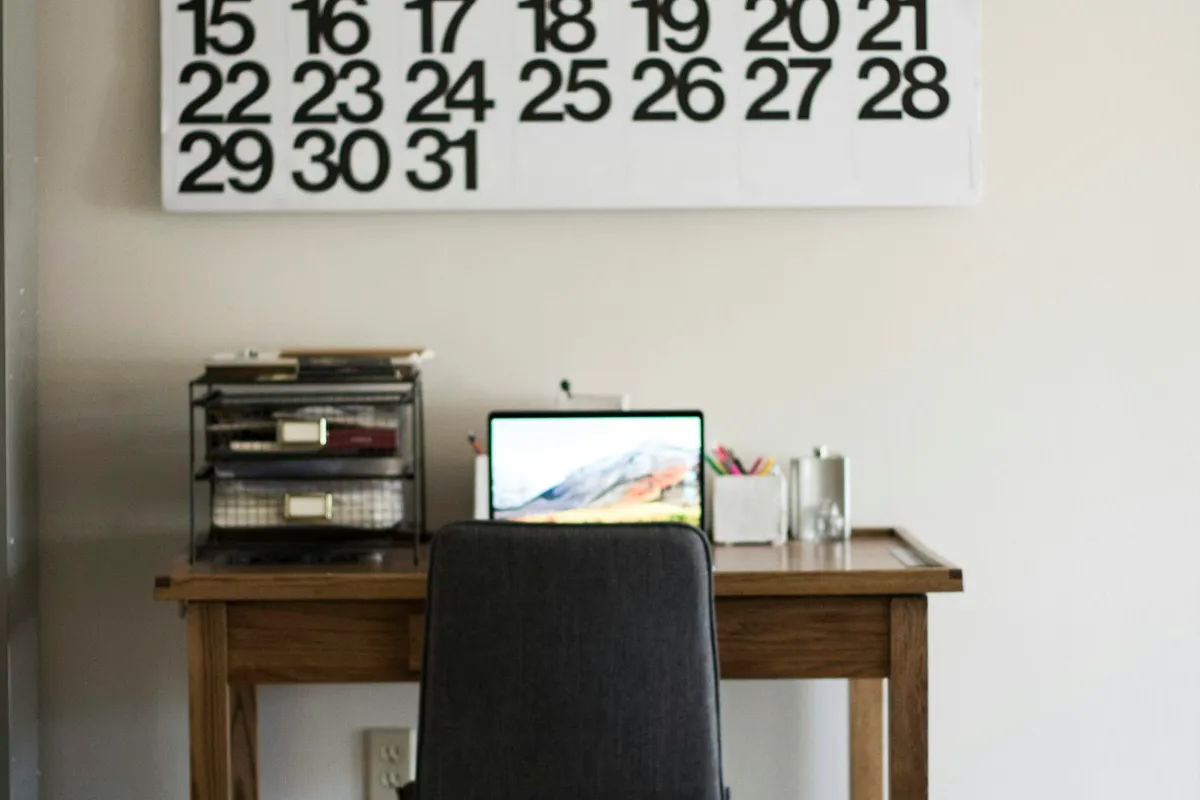 A large wall calendar ending on the 31st hangs above a small wooden desk with an open laptop, a wire paper tray, a cup of colored pens and an empty desk chair