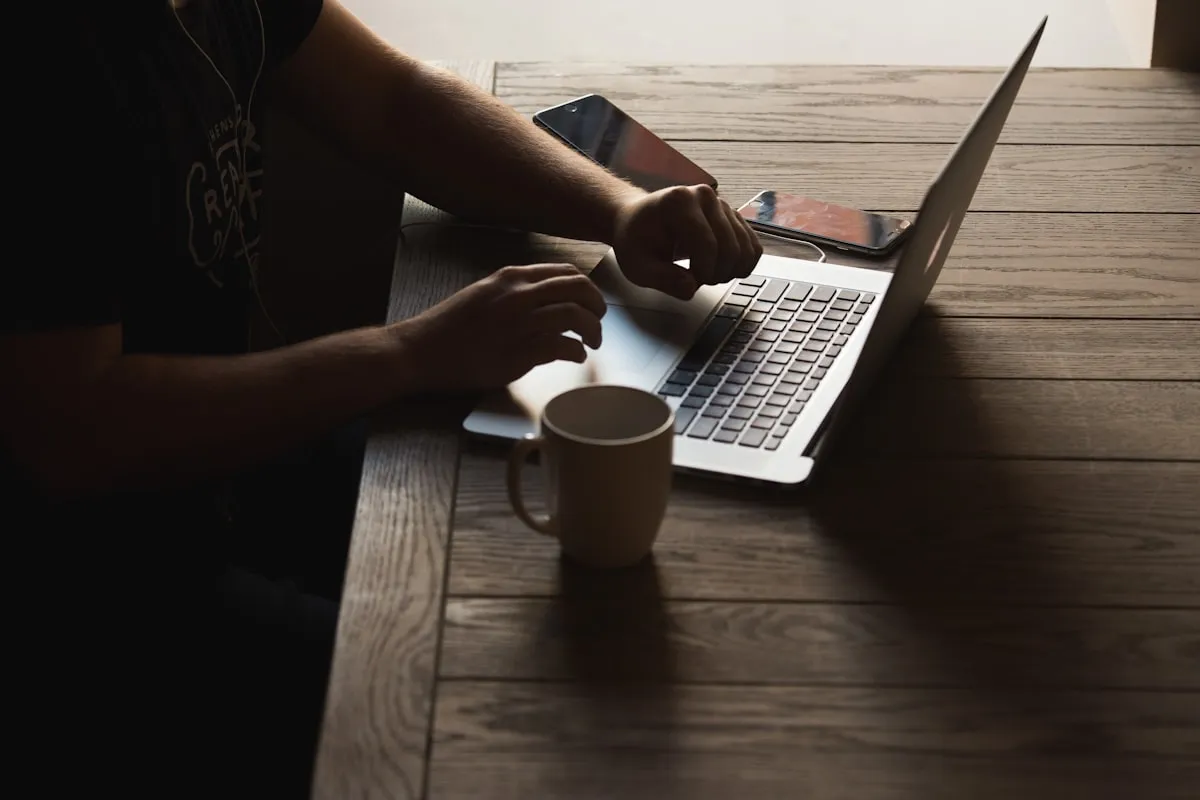 A person working on a laptop at a wooden desk with a coffee mug and phone beside them